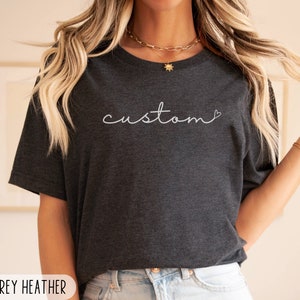 May include: Dark grey heather t-shirt with the word "custom" in white script. The Simple West logo is in the upper right corner. The shirt is a crew neck style.