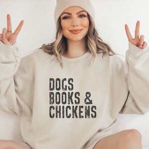 May include: A beige sweatshirt with the text "DOGS BOOKS & CHICKENS" printed in black on the front. The sweatshirt is being worn by a person with a light brown beanie.