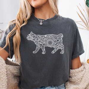 May include: Dark gray t-shirt with a white decorative cat design. The cat is filled with floral and geometric patterns. The text "MEDIUM PEPPER TEE" is at the bottom of the shirt. The Simple West logo is in the upper right corner.