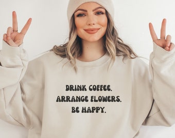 May include: A woman wearing a light beige sweatshirt with the text "Drink coffee, arrange flowers, be happy." printed on the front.