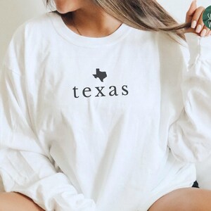 May include: A white long-sleeve tee with the word "texas" printed in black, along with a black outline of the state. The shirt is a large size and is made of a soft material. The Simple West logo is visible in the upper right corner.