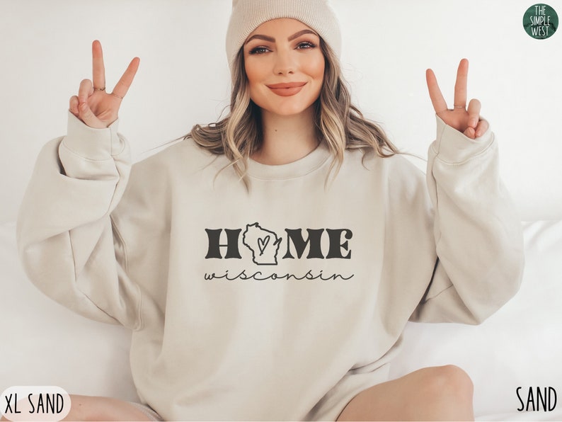 May include: A beige sweatshirt with the text "HOME wisconsin" and a graphic of the state of Wisconsin with a heart inside. The sweatshirt is being modeled by a person wearing a beige beanie.