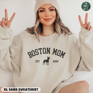 Puede incluir: Sudadera de color arena con el texto "BOSTON MOM" en negrita, en forma de arco, con "EST 2025" y una silueta de perro. La sudadera es de color neutro y tiene cuello redondo. Las palabras "XL SAND SWEATSHIRT" est&aacute;n en la parte inferior.