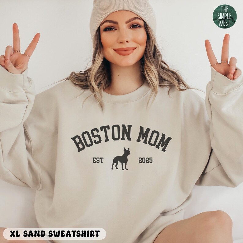 Puede incluir: Sudadera de color arena con el texto "BOSTON MOM" en negrita, en forma de arco, con "EST 2025" y una silueta de perro. La sudadera es de color neutro y tiene cuello redondo. Las palabras "XL SAND SWEATSHIRT" est&aacute;n en la parte inferior.