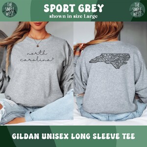 May include: Sport grey Gildan unisex long sleeve tee. The front of the shirt has the words "north carolina" in a script font. The back of the shirt has a black graphic of the North Carolina state outline with a decorative pattern.