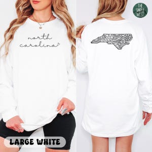 May include: White long-sleeve shirt with "north carolina" in cursive on the front. The back features a black floral design in the shape of the state. The Simple West logo is visible. The shirt is labeled "LARGE WHITE".