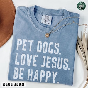 May include: Blue jean colored t-shirt with the text "PET DOGS. LOVE JESUS. BE HAPPY." in white block letters. A gold necklace with a pendant rests on the shirt. The shirt is a Comfort Colors brand.