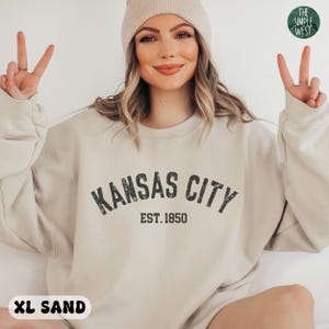 K&ouml;nnte beinhalten: Ein beiger Pullover mit dem Schriftzug "Kansas City Est. 1850" in Schwarz. Der Pullover ist locker geschnitten und hat einen Rundhalsausschnitt.