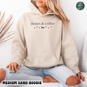 May include: A medium sand-colored hoodie with a front pocket and drawstring hood. The text "doxies & coffee" is printed on the front, along with a dachshund silhouette and paw prints. The words "MEDIUM SAND HOODIE" are printed at the bottom.
