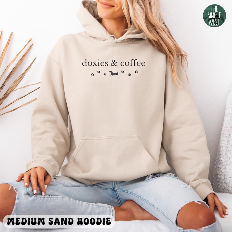 May include: A medium sand-colored hoodie with a front pocket and drawstring hood. The text "doxies & coffee" is printed on the front, along with a dachshund silhouette and paw prints. The words "MEDIUM SAND HOODIE" are printed at the bottom.