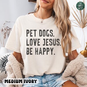 Puede incluir: Camiseta de color marfil medio con el texto "PET DOGS. LOVE JESUS. BE HAPPY." en letras negras. El logotipo de The Simple West est&aacute; en la esquina superior derecha.