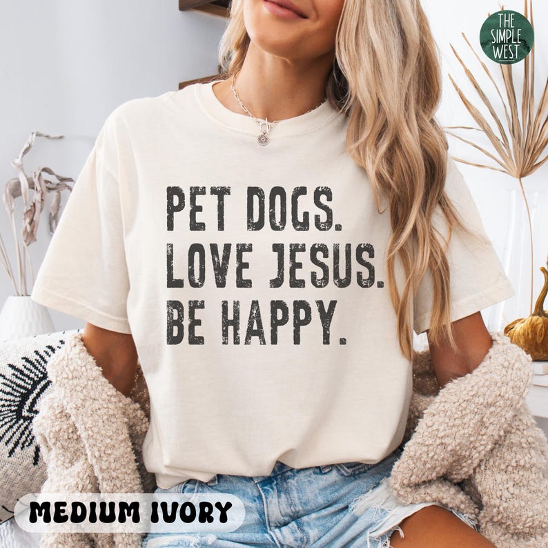 Puede incluir: Camiseta de color marfil medio con el texto "PET DOGS. LOVE JESUS. BE HAPPY." en letras negras. El logotipo de The Simple West est&aacute; en la esquina superior derecha.