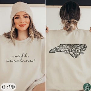 May include: A beige sweatshirt with the text "north carolina" on the front and a black and white mandala design of the state of North Carolina on the back. The sweatshirt is available in size XL and the colour sand.