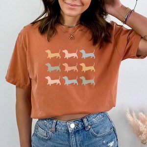 May include: A rust-coloured t-shirt with a graphic of nine pastel-coloured dachshund silhouettes. The text "YAM" is in the top left corner of the image.