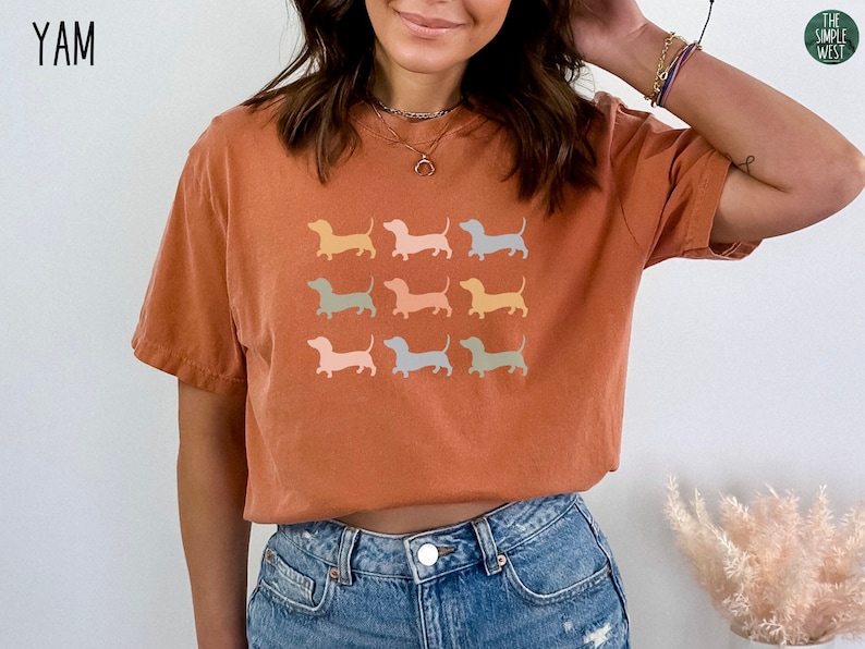 May include: A rust-coloured t-shirt with a graphic of nine pastel-coloured dachshund silhouettes. The text "YAM" is in the top left corner of the image.