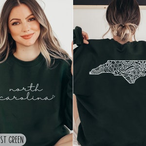 May include: Forest green sweatshirt with a white outline of the state of North Carolina featuring a floral design. The front of the sweatshirt has the text "north carolina" in white cursive writing with a heart.