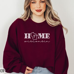 May include: A maroon sweatshirt with the text "HOME" and a heart-shaped outline of the state of Wisconsin. The text "Wisconsin" is written in cursive below the state outline. The sweatshirt is available in size 2XL.