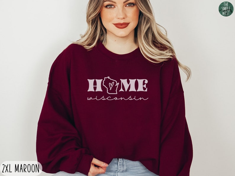 May include: A maroon sweatshirt with the text "HOME" and a heart-shaped outline of the state of Wisconsin. The text "Wisconsin" is written in cursive below the state outline. The sweatshirt is available in size 2XL.