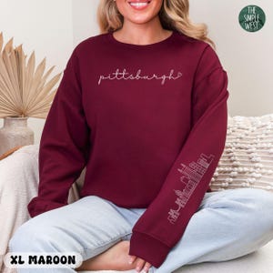 Op de afbeelding: Bordeaux sweatshirt met het woord "pittsburgh" in cursiefschrift over de borst. De rechter mouw heeft een witte omtrek van een skyline. Het Simple West-logo staat rechtsboven. Maat XL Maroon.