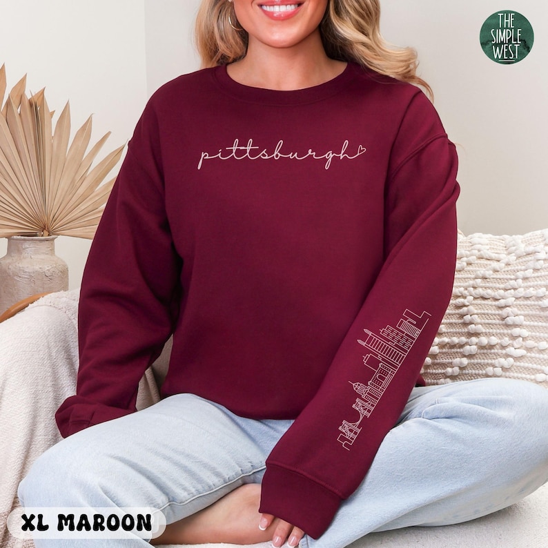 Op de afbeelding: Bordeaux sweatshirt met het woord "pittsburgh" in cursiefschrift over de borst. De rechter mouw heeft een witte omtrek van een skyline. Het Simple West-logo staat rechtsboven. Maat XL Maroon.
