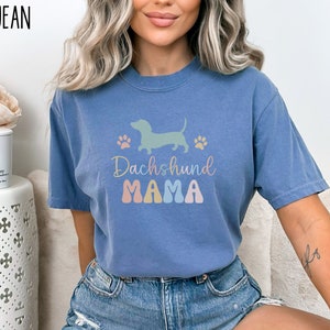 Puede incluir: Una camiseta azul jean con una silueta de dachshund pastel y el texto "Dachshund Mama" en colores pastel.