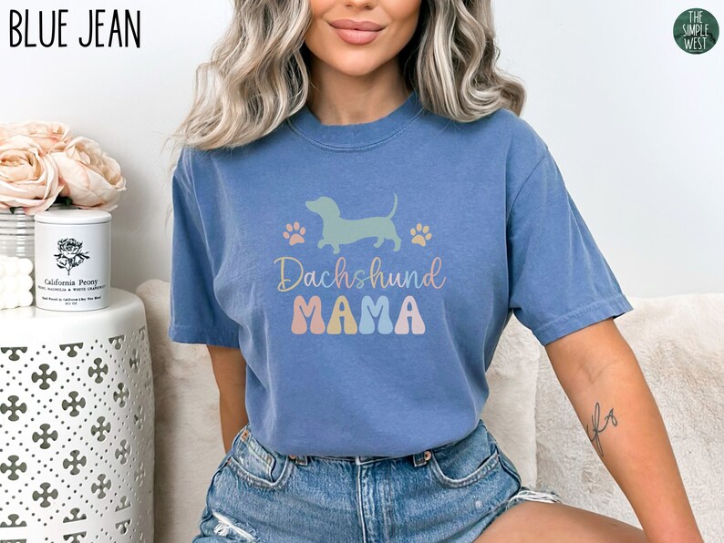 Puede incluir: Una camiseta azul jean con una silueta de dachshund pastel y el texto "Dachshund Mama" en colores pastel.