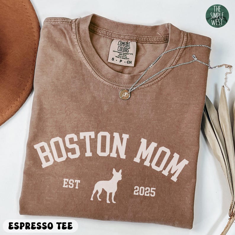 Puede incluir: Camiseta color espresso con el texto "BOSTON MOM" en letras blancas, una silueta de perro y el a&ntilde;o "2025". La camiseta tambi&eacute;n tiene el texto "EST" y "ESPRESSO TEE". Un collar plateado con un colgante descansa sobre la camiseta.