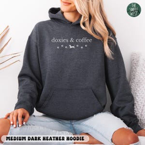 May include: Medium dark heather hoodie with a front pocket and a drawstring hood. The hoodie features the text "doxies & coffee" in white, along with paw prints and a dachshund illustration. The bottom of the hoodie reads "MEDIUM DARK HEATHER HOODIE".