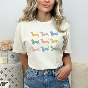 May include: A woman is wearing a light ivory t-shirt with a pattern of nine pastel coloured dachshund silhouettes. The shirt is a medium size.