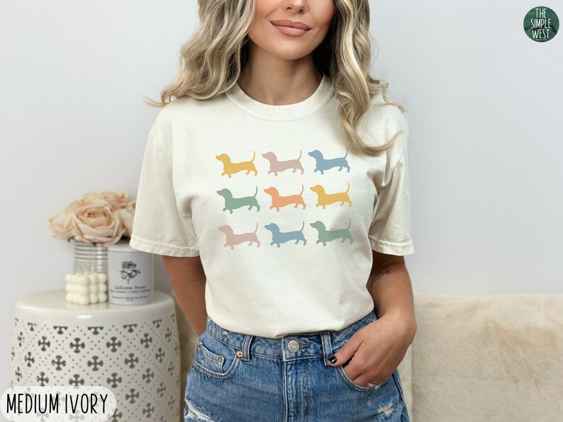 May include: A woman is wearing a light ivory t-shirt with a pattern of nine pastel coloured dachshund silhouettes. The shirt is a medium size.