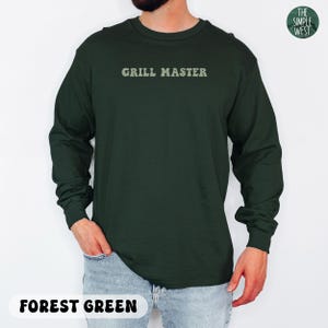 May include: Forest green long-sleeve shirt with the text "GRILL MASTER" in a light green font. The shirt is a solid color and has a crew neck. The Simple West logo is in the upper right corner.