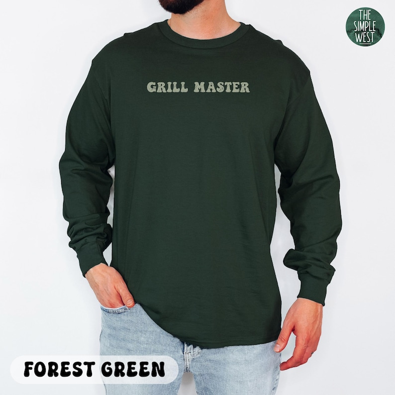 May include: Forest green long-sleeve shirt with the text "GRILL MASTER" in a light green font. The shirt is a solid color and has a crew neck. The Simple West logo is in the upper right corner.