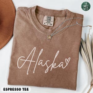 May include: Espresso-colored t-shirt with the word "Alaska" in white script and a small heart. The shirt has a crew neck and a Comfort Colors tag. A silver necklace with a pendant is draped across the shirt. The Simple West logo is in the upper right corner.