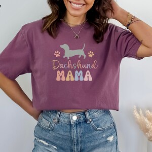 Puede incluir: Una camiseta corta de color malva con un gr&aacute;fico de un dachshund y el texto "Dachshund Mama" en letras de colores.