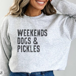Op de afbeelding: Een grijs sweatshirt met de tekst "Weekends Dogs & Pickles" in zwart gedrukt.