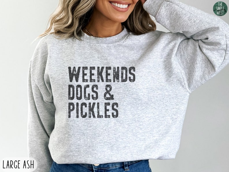 Op de afbeelding: Een grijs sweatshirt met de tekst "Weekends Dogs & Pickles" in zwart gedrukt.