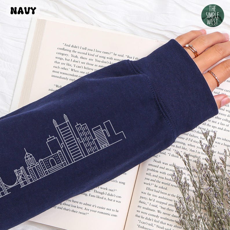 Op de afbeelding: Marineblauwe longsleeve met een witte skyline-afbeelding. Het shirt heeft het woord "NAVY" linksboven. Het Simple West-logo staat rechtsboven. Het shirt is gemaakt van een zacht, comfortabel materiaal.