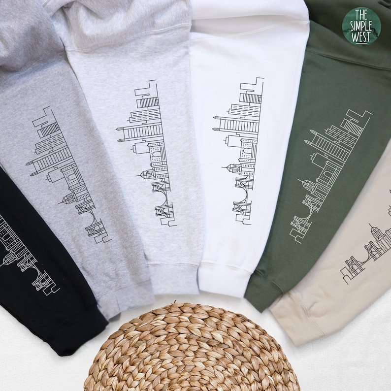 Op de afbeelding: Een collectie sweatshirts in diverse kleuren, waaronder zwart, grijs, wit en groen. Elke mouw heeft een zwart-witte lijntekening van een skyline. Het logo The Simple West is zichtbaar.