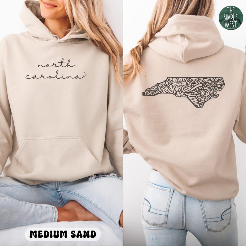 Op de afbeelding: Een beige hoodie met de woorden "north carolina" in cursief op de voorkant. De achterkant heeft een zwart, patroon van North Carolina. De hoodie heeft een voorzak en een capuchon. De tekst "MEDIUM SAND" staat onderaan.