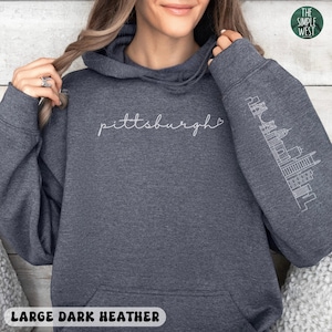 Op de afbeelding: Donkergrijze hoodie met het woord "Pittsburgh" in wit script over de borst. De rechter mouw heeft een witte omtrek van de skyline van Pittsburgh. De hoodie heeft een capuchon met trekkoord en een voorzak. De tekst "LARGE DARK HEATHER" staat onderaan.