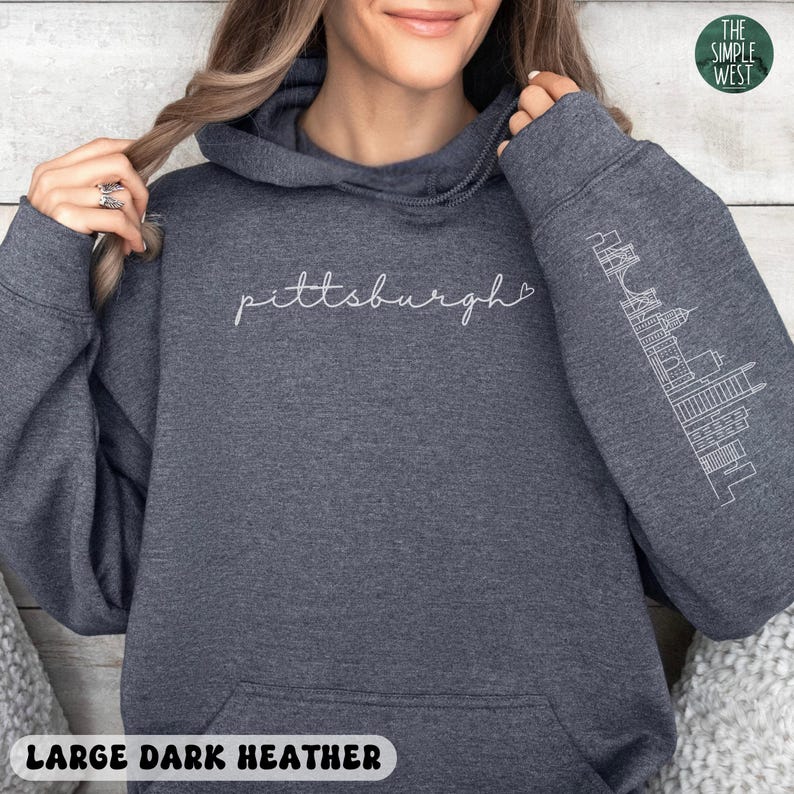 Op de afbeelding: Donkergrijze hoodie met het woord "Pittsburgh" in wit script over de borst. De rechter mouw heeft een witte omtrek van de skyline van Pittsburgh. De hoodie heeft een capuchon met trekkoord en een voorzak. De tekst "LARGE DARK HEATHER" staat onderaan.