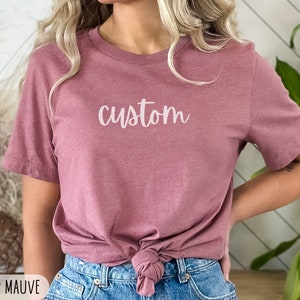以下が含まれることがあります： 白の筆記体で「custom」と書かれた、ヘザーモーヴのTシャツ。シャツはウエストで結ばれ、ブルージーンズと合わせています。The Simple Westのロゴが右上に見えます。