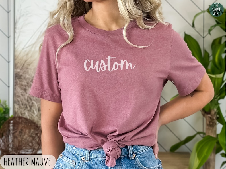 以下が含まれることがあります： 白の筆記体で「custom」と書かれた、ヘザーモーヴのTシャツ。シャツはウエストで結ばれ、ブルージーンズと合わせています。The Simple Westのロゴが右上に見えます。