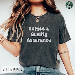 May include: A dark gray t-shirt with the text "Coffee & Quality Assurance" in white, retro-style lettering. The shirt is being worn by a person with long blonde hair.