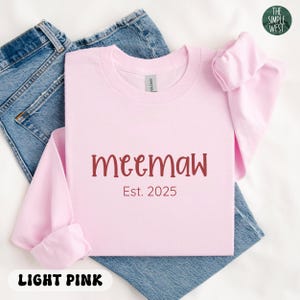 May include: A light pink sweatshirt with the word "meemaw" in maroon script and the text "Est. 2025". The sweatshirt is folded on top of a pair of blue jeans. The words "Light Pink" are at the bottom.