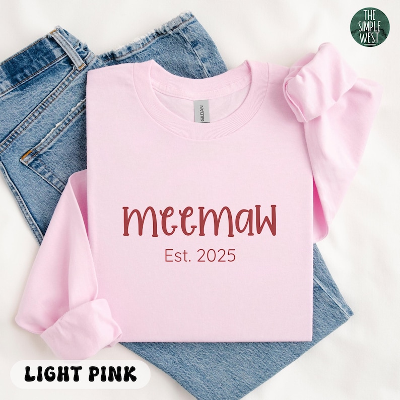 May include: A light pink sweatshirt with the word "meemaw" in maroon script and the text "Est. 2025". The sweatshirt is folded on top of a pair of blue jeans. The words "Light Pink" are at the bottom.