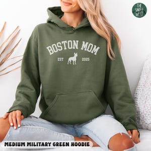 Puede incluir: Sudadera con capucha verde militar mediana con bolsillo de canguro y capucha con cord&oacute;n. El texto blanco en la parte delantera dice "BOSTON MOM" encima de una silueta de perro, con "EST" y "2025" debajo. El logotipo de The Simple West est&aacute; en la esquina superior derecha.