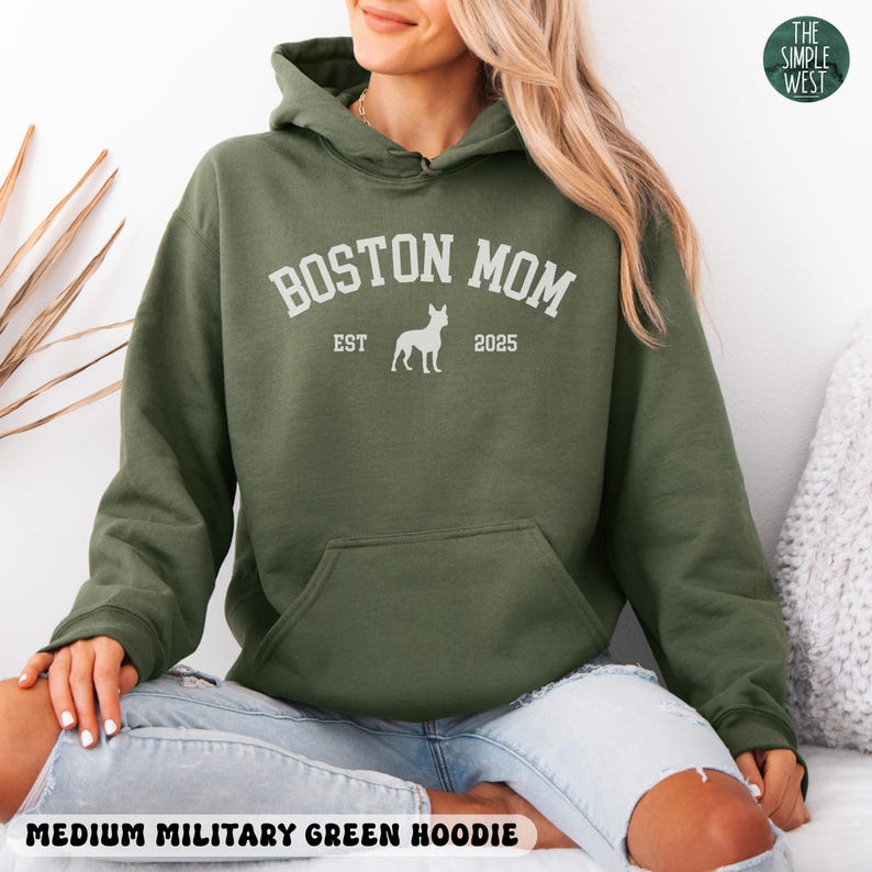 Puede incluir: Sudadera con capucha verde militar mediana con bolsillo de canguro y capucha con cord&oacute;n. El texto blanco en la parte delantera dice "BOSTON MOM" encima de una silueta de perro, con "EST" y "2025" debajo. El logotipo de The Simple West est&aacute; en la esquina superior derecha.