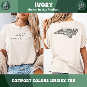 Pu&ograve; includere: T-shirt unisex Comfort Colors avorio. La parte anteriore presenta la scritta "north carolina" in corsivo. Il retro mostra un design nero dettagliato del contorno dello stato della Carolina del Nord. Il logo The Simple West &egrave; visibile.
