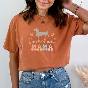Puede incluir: Una mujer lleva una camiseta naranja quemada con una silueta de dachshund y el texto "Dachshund Mama" en colores pastel.